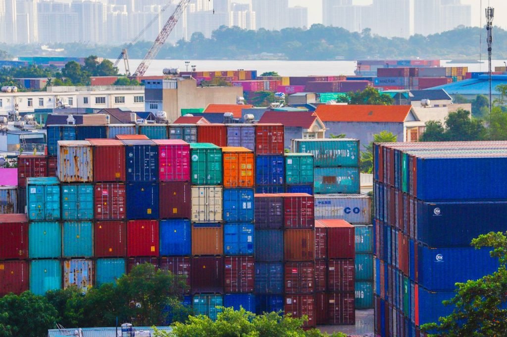 Vibrant stacked cargo containers at a bustling port with urban skyline in background.